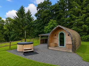 A wooden cabin with a curved roof, a small porch, and a hot tub in a forested area.