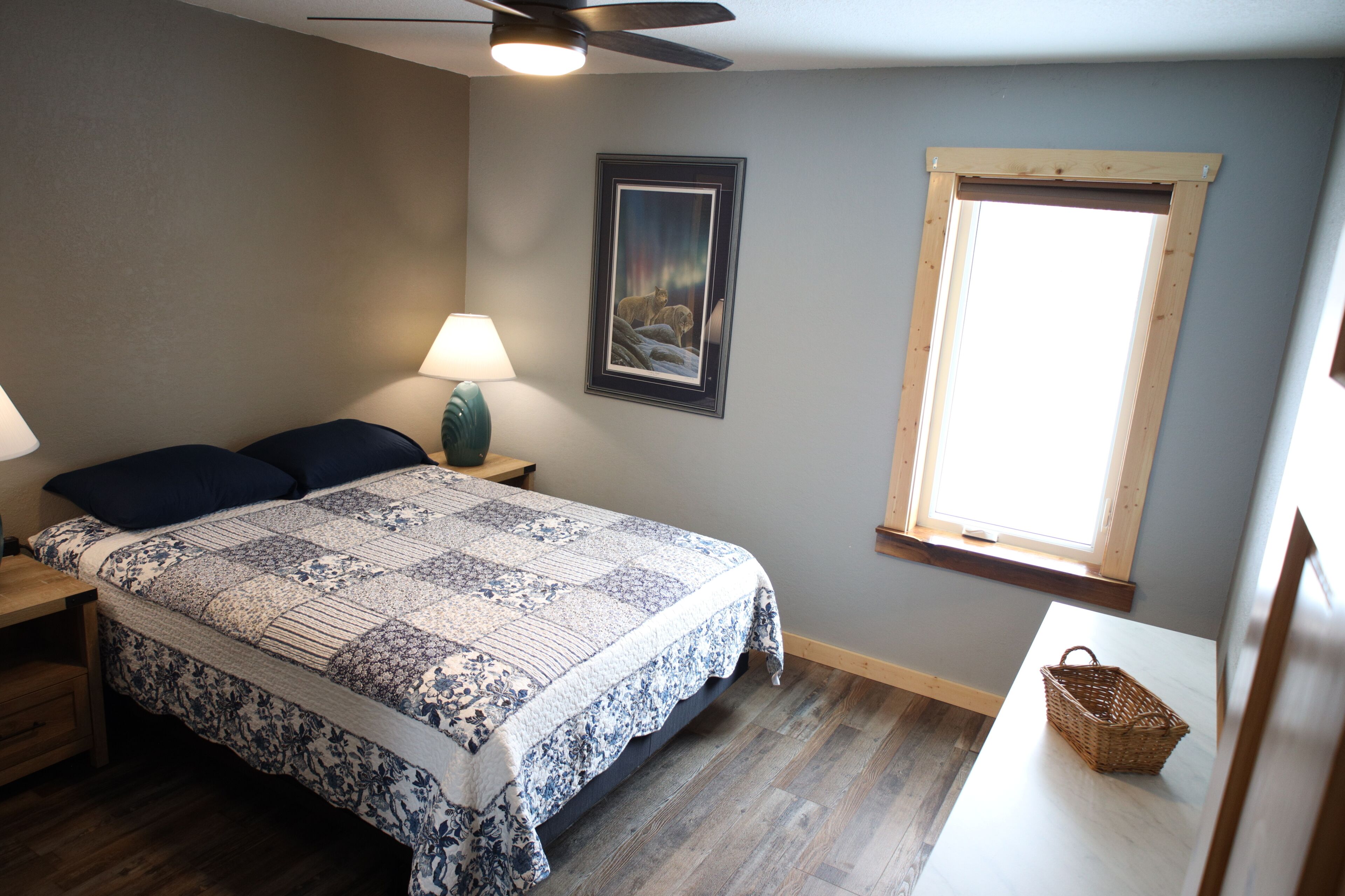 A bedroom with a bed, a nightstand, a lamp, a framed picture, a window, and a wooden floor.