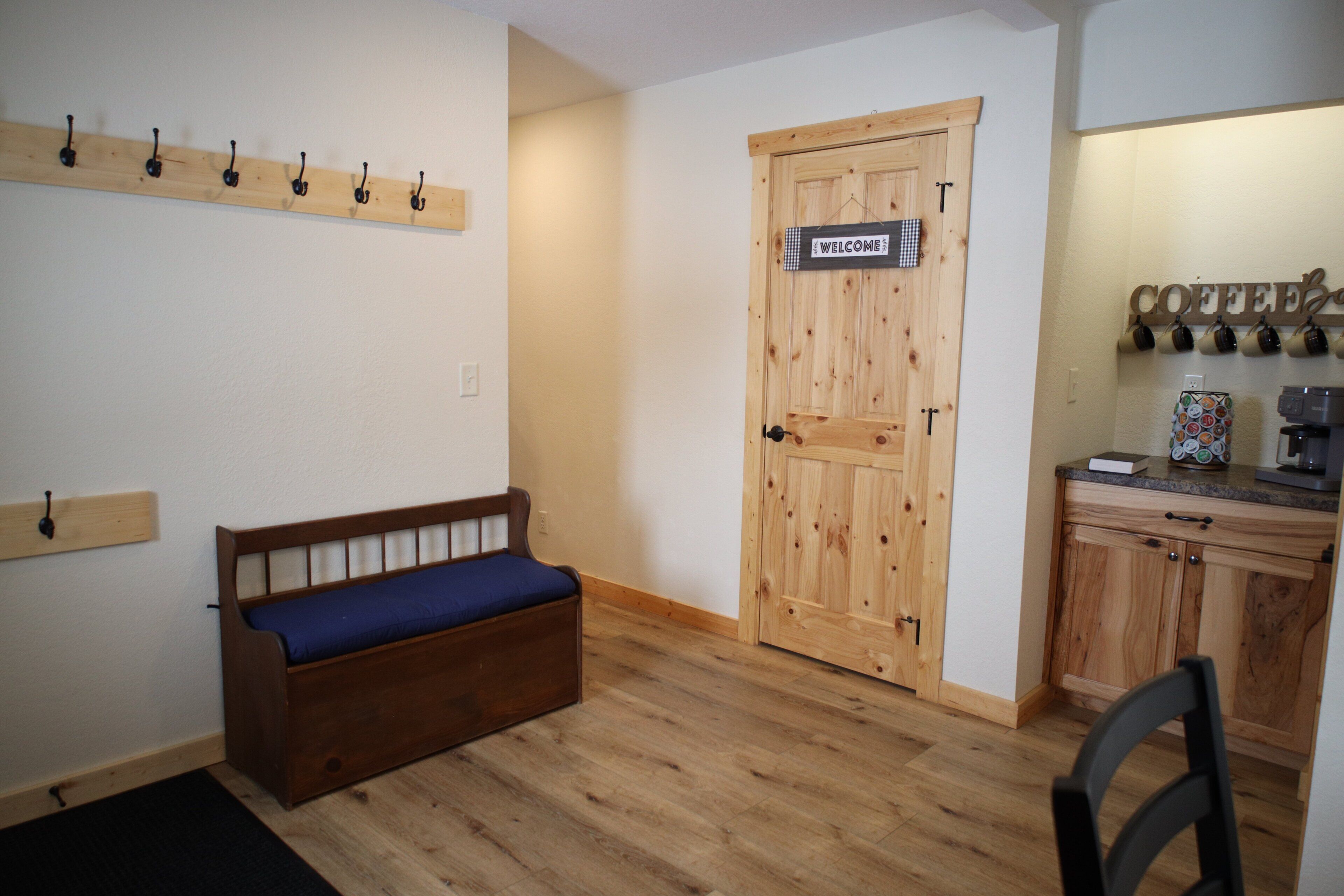 Une pièce avec un banc en bois, une porte en bois ornée d’une pancarte de bienvenue, un coin café et une petite table.