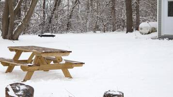 Une table de pique-nique en bois et un foyer, installés dans un décor extérieur enneigé, avec des arbres et un bâtiment en arrière-plan.