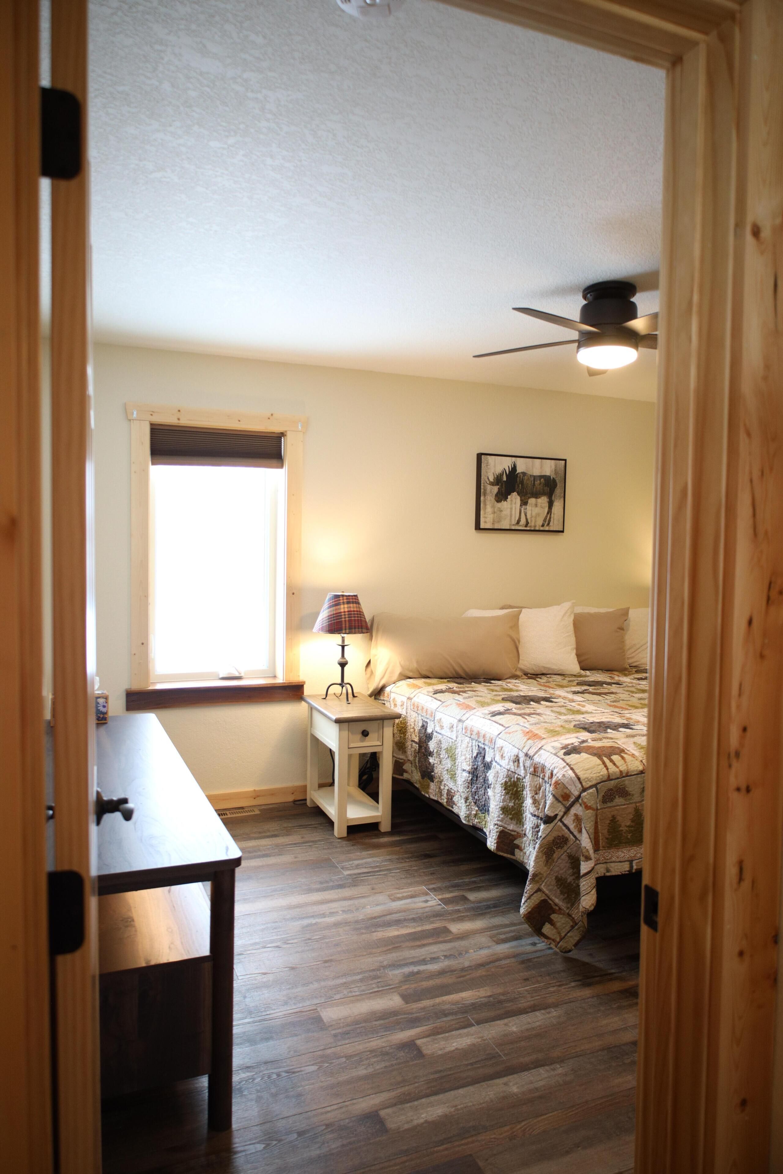 Une chambre à coucher avec un lit, une table de chevet, une lampe, un ventilateur de plafond et une fenêtre.