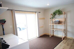 A laundry room with a washing machine, a broom, a sliding glass door, and a wooden shelf with a plant and basket.