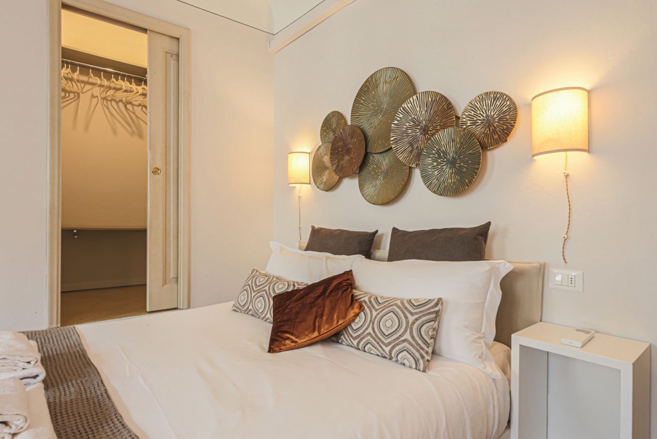 Un lit bien fait, agrémenté d’oreillers à motifs, une table de chevet avec une lampe, et un mur orné d’éléments décoratifs circulaires.