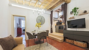 A living room with a sofa, dining table, chairs, a clock, a TV, and a staircase leading to a kitchen. - Apartments Florence - Mezzo Boutique Vista (Firenze)