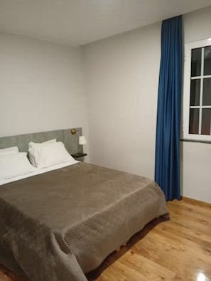 Une chambre à coucher avec un lit, une table de chevet, une lampe, une fenêtre avec des rideaux et un plancher en bois.
