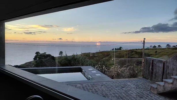 A modern room with a large window overlooking a coastal landscape at sunset. - Casas do Pargo - Oak Tree House (Lombada Velha)