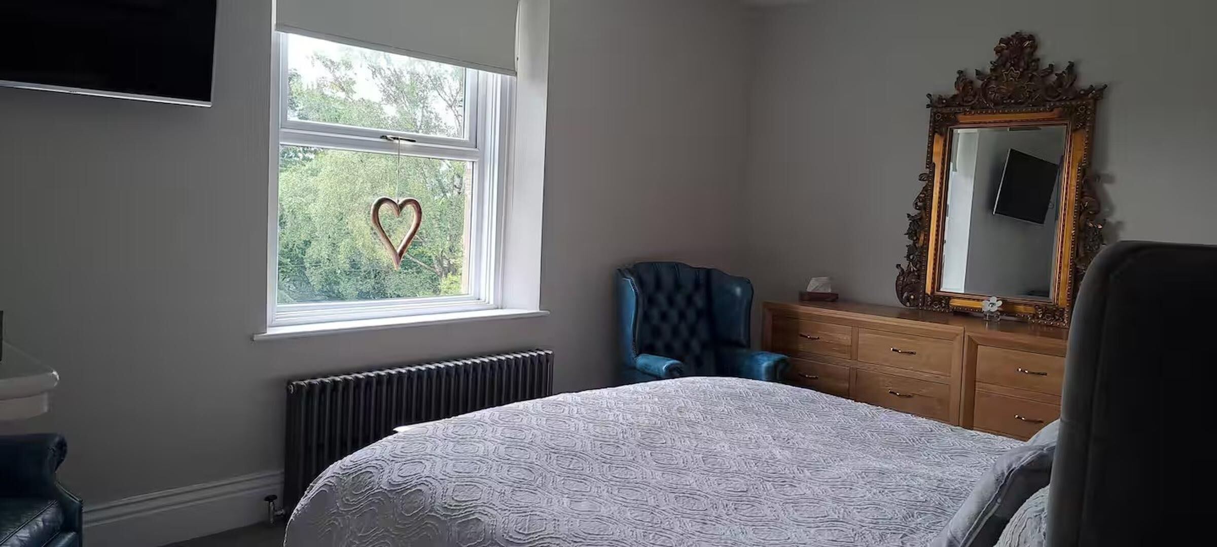 A bedroom with a bed, a dresser, a chair, a mirror, and a window with a heart decoration.