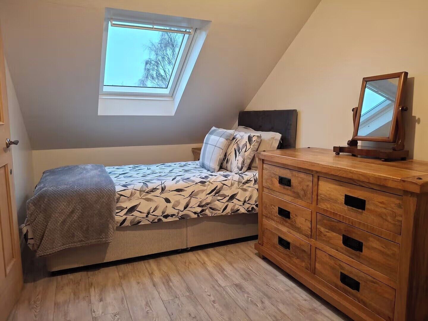A bedroom with a sloped ceiling, a skylight, a bed with patterned bedding, a wooden dresser, and a mirror.