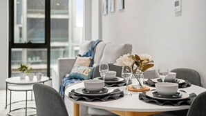 A dining area with a round table set for four, white plates, black placemats, and a vase with white flowers. - BNBIFY Stylish Melbourne CBD 2BR Apartment | Pool, Spa, Gym (Melbourne)
