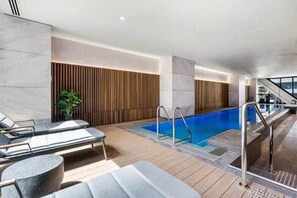 A modern indoor pool area with lounge chairs, a wooden wall, and a staircase. - BNBIFY Stylish Melbourne CBD 2BR Apartment | Pool, Spa, Gym (Melbourne)