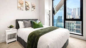 A modern bedroom with a large bed, a nightstand, a potted plant, and a city view through a large window. - BNBIFY Stylish Melbourne CBD 2BR Apartment | Pool, Spa, Gym (Melbourne)