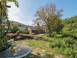 Eine ländliche Szene im Freien mit einem Steintisch, einer Flasche und einem Glas, umgeben von viel Grün und einem Gebäude mit geneigtem Dach.
