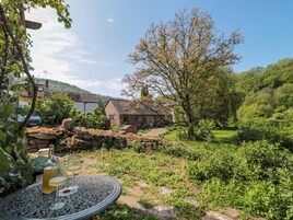 Entorno rústico al aire libre con una mesa de piedra, una botella y una copa, rodeado de follaje y un edificio con techo inclinado.