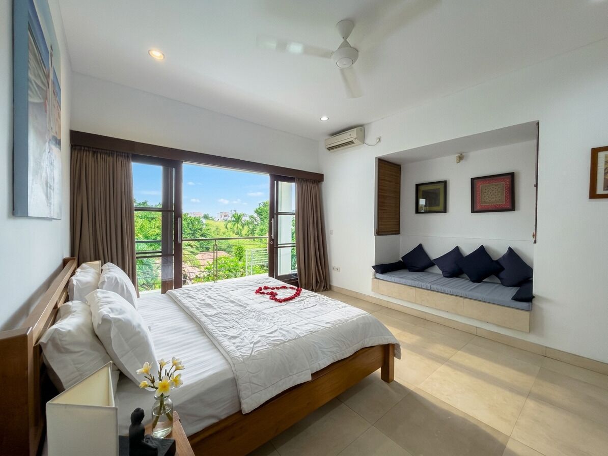 Un dormitorio con cama, ventana con cortinas, ventilador de techo y asiento con cojines sujetos a la pared.