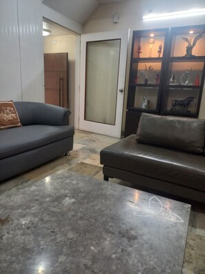 A living room with a grey sofa, a smaller grey sofa, a coffee table with a geometric design, a door with frosted glass, and a shelf with decorative items.
