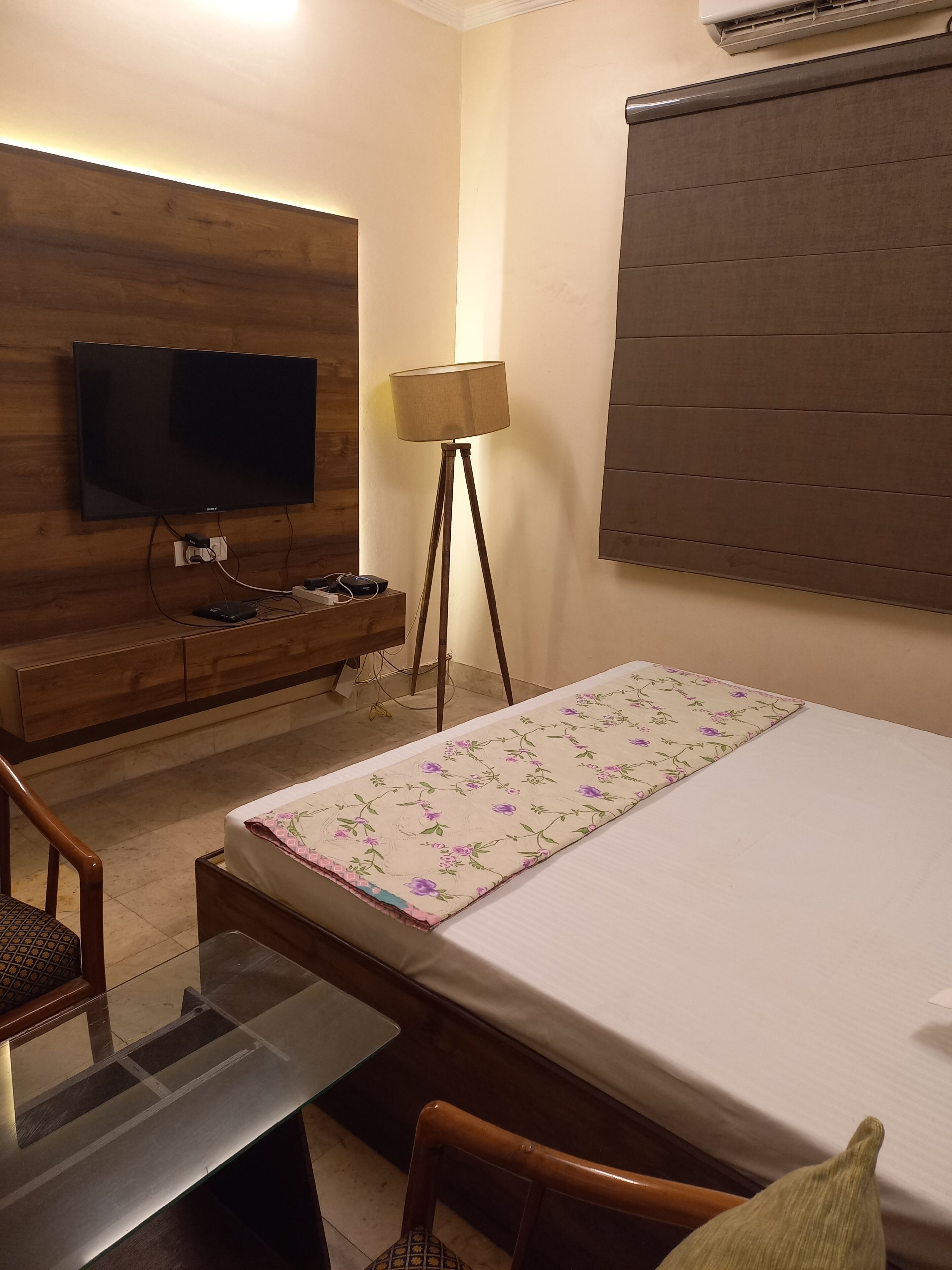 Een hotelkamer met een bed, een televisie aan een houten wand bevestigd, een vloerlamp en een raam met zonwering.
