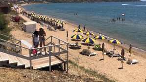 En strand med en trätrappa som leder ner till sanden, rader av strandparasoller och människor som njuter av vattnet.