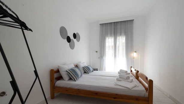 A bedroom with a wooden bed, two pillows, a black and white patterned rug, a wall-mounted light, and a window with curtains. - Erato Apartment 1 by RentalsPro - Nea Plagia Halkidiki (Plagia)