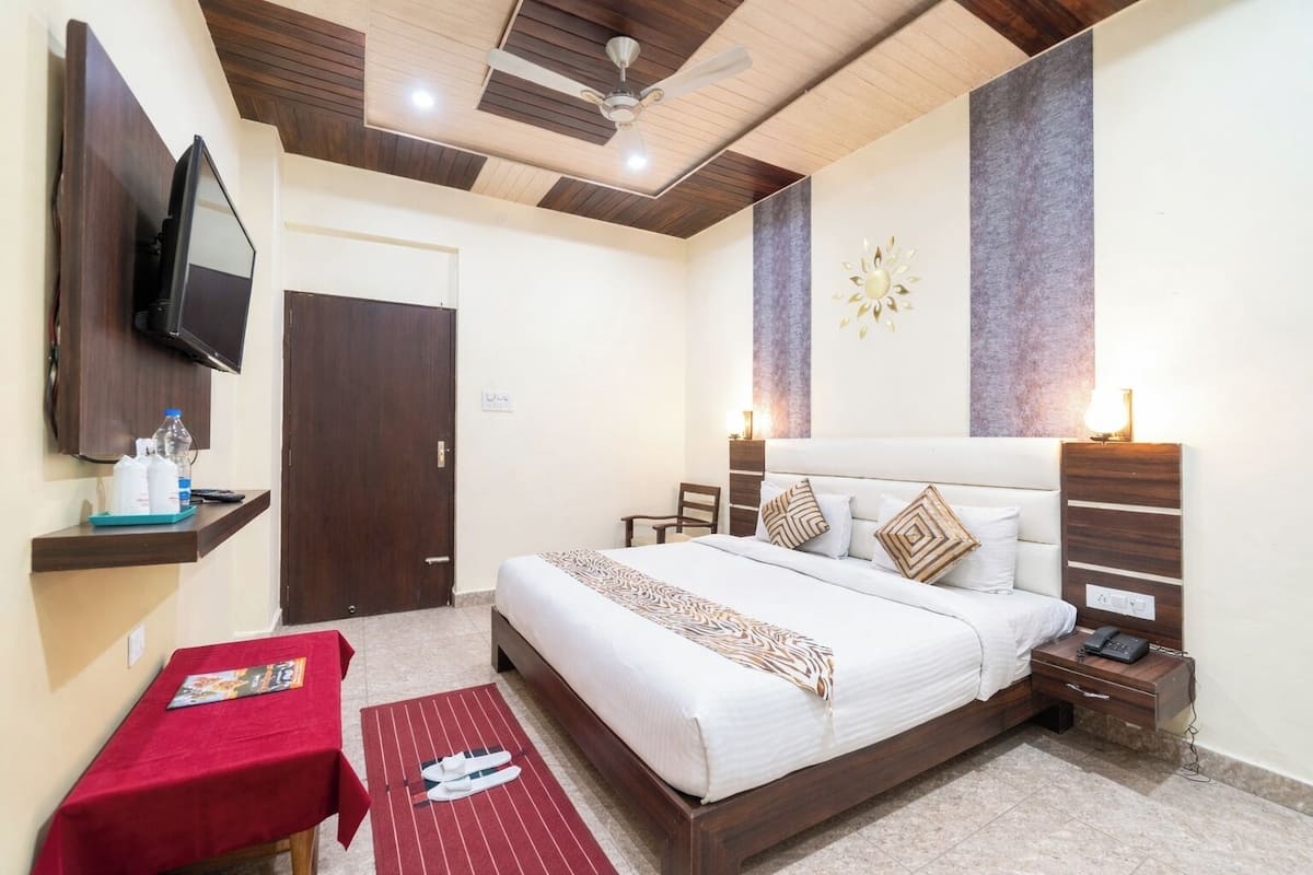 Habitación de hotel con cama, televisor montado en la pared, mesita de noche con lámpara, una mesa roja con una revista y un tapete rojo con zapatillas.