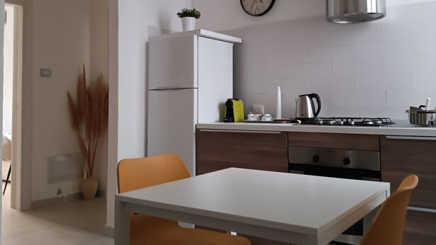 A modern kitchen with a white refrigerator, stove, and stainless steel range hood. There is a dining table with two chairs and a potted plant. - YOUR HOUSE (Olbia)
