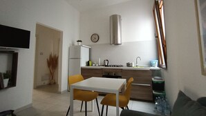 A compact kitchen with a refrigerator, stove, and sink. There is a dining table with chairs and a sofa in the living area. - YOUR HOUSE (Olbia)