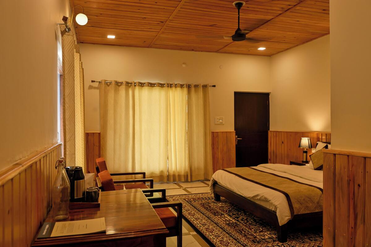 A hotel room with a bed, a desk with a chair, a small table with a bottle, a lamp, and a door.