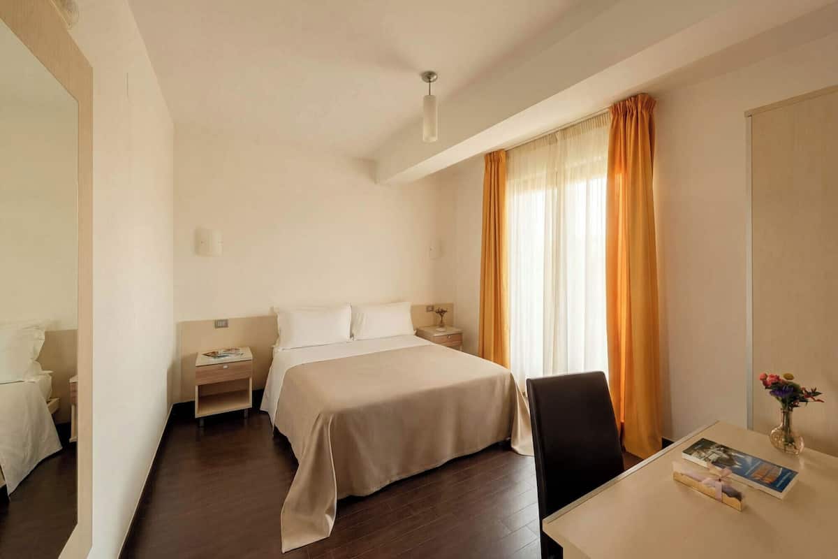 Une chambre d’hôtel comprenant un lit, des tables de chevet, un bureau avec une chaise et une fenêtre avec des rideaux.