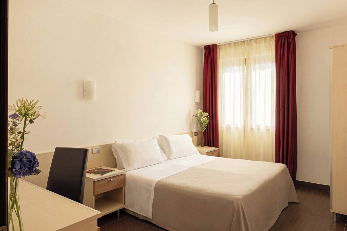 Une chambre d’hôtel avec un lit, un bureau, une chaise, une table de chevet et une fenêtre avec des rideaux.
