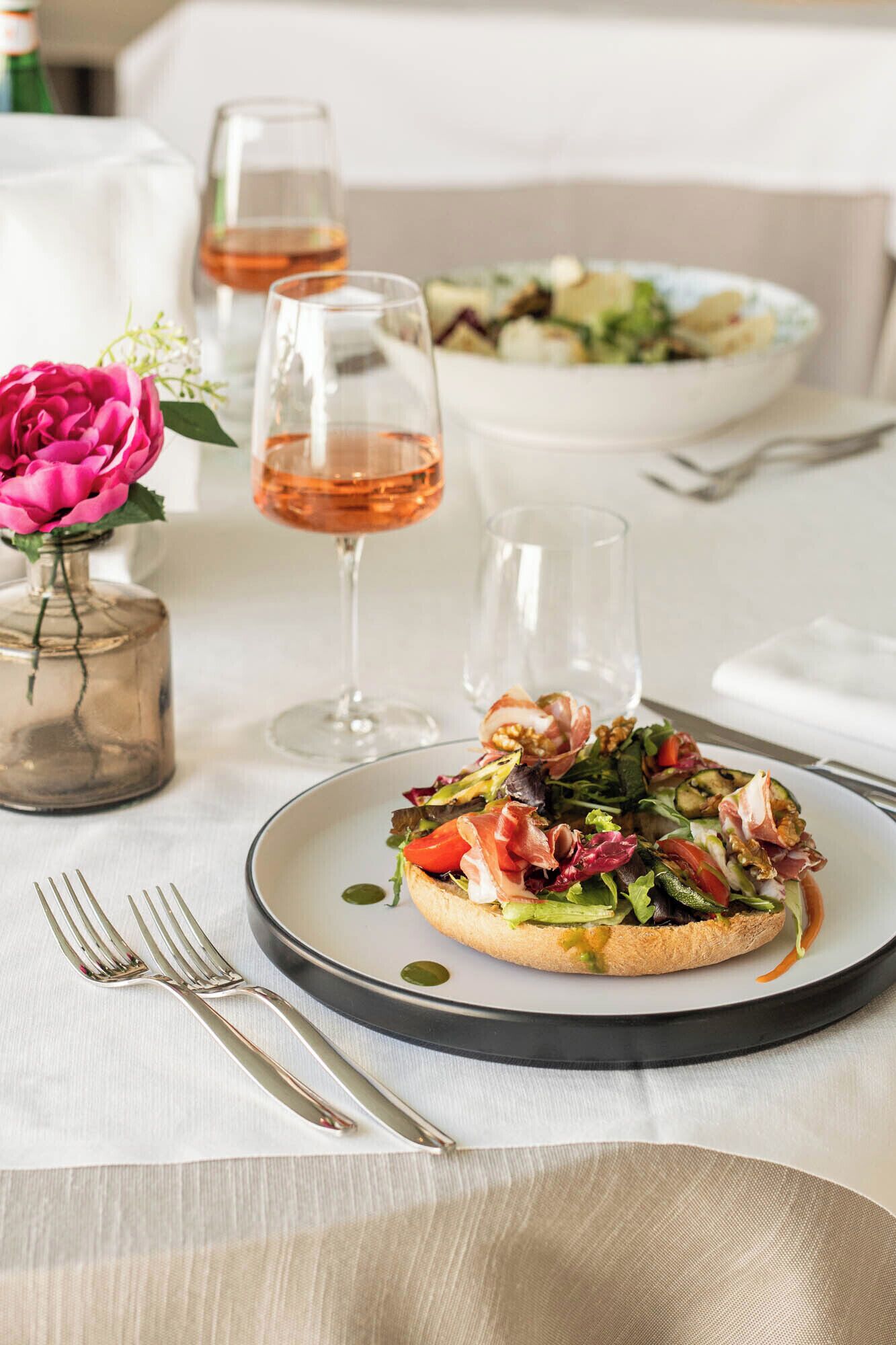 Une table à manger avec une assiette de nourriture, deux verres à vin et un vase avec une fleur.