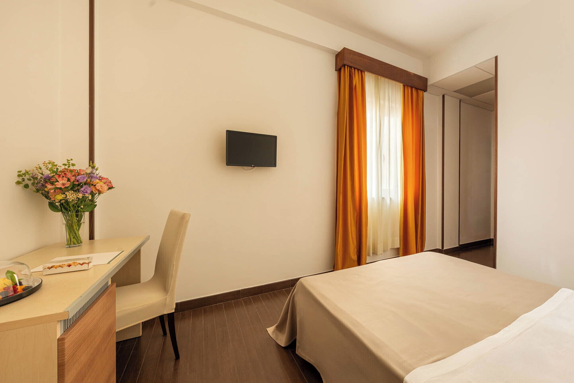 A hotel room with a bed, a desk with a chair, a TV, and a window with curtains.