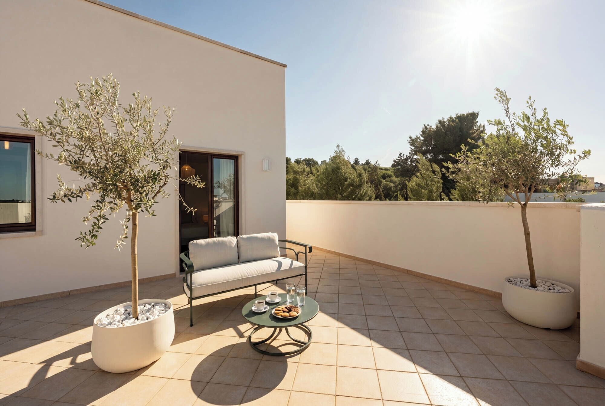 Un espace extérieur moderne, aménagé avec un canapé, une table ronde sur laquelle sont disposées des boissons et deux arbres en pot.