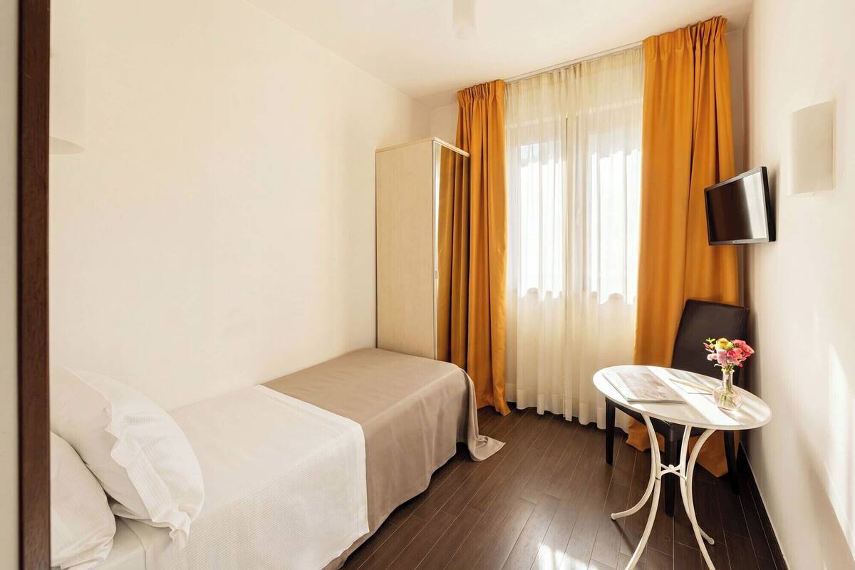 Une petite chambre d’hôtel, bien rangée, avec un lit, une table sur laquelle est posé un vase, une chaise, un téléviseur et une fenêtre avec des rideaux.