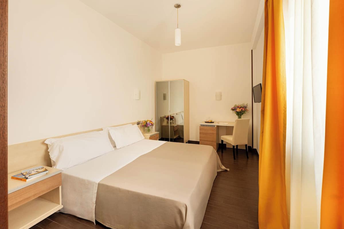 Une chambre d’hôtel comprenant un lit, une table de chevet, un miroir, un bureau, une chaise et une fenêtre avec des rideaux.