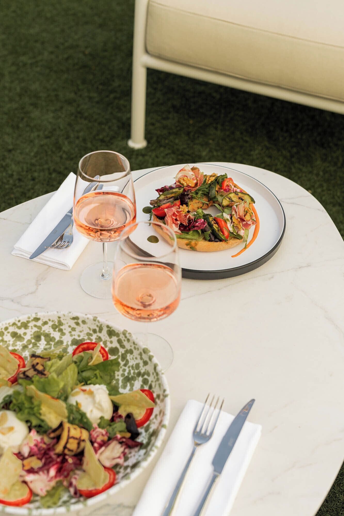 A table with two plates of food and two glasses of wine, set on a marble table outdoors.