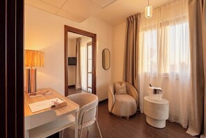 A hotel room with a desk, two chairs, a round coffee table, a mirror, and a window with curtains. - ARBORIS BELLI HOTEL  (Alberobello)