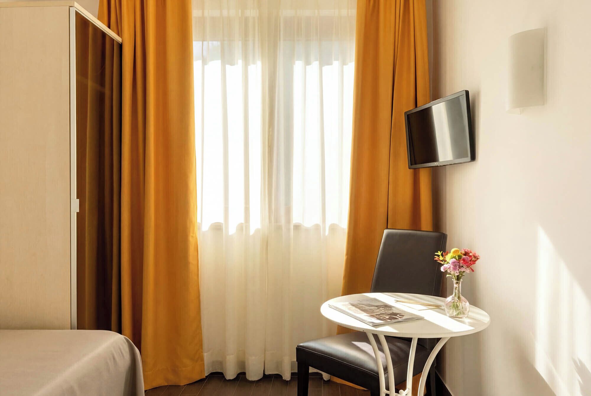 A hotel room with a bed, a table with a vase of flowers, a chair, and a TV mounted on the wall.