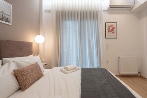 A neatly made bed with white linens, a brown accent pillow, and a folded white towel. A gray throw blanket is draped over the bed. The room features a large window with sheer curtains, a wall-mounted air conditioner, and a framed picture of a flower. - THE EDGE Luxurious Penthouse Shared Gym (Kallithea)