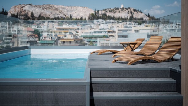 A rooftop pool area with wooden loungers and a view of a cityscape with a prominent rocky hill in the background. - THE EDGE Luxurious Penthouse Shared Gym (Kallithea)