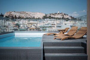 Un área de piscina en la azotea con tumbonas de madera y vistas a un paisaje urbano con una colina rocosa destacada al fondo.