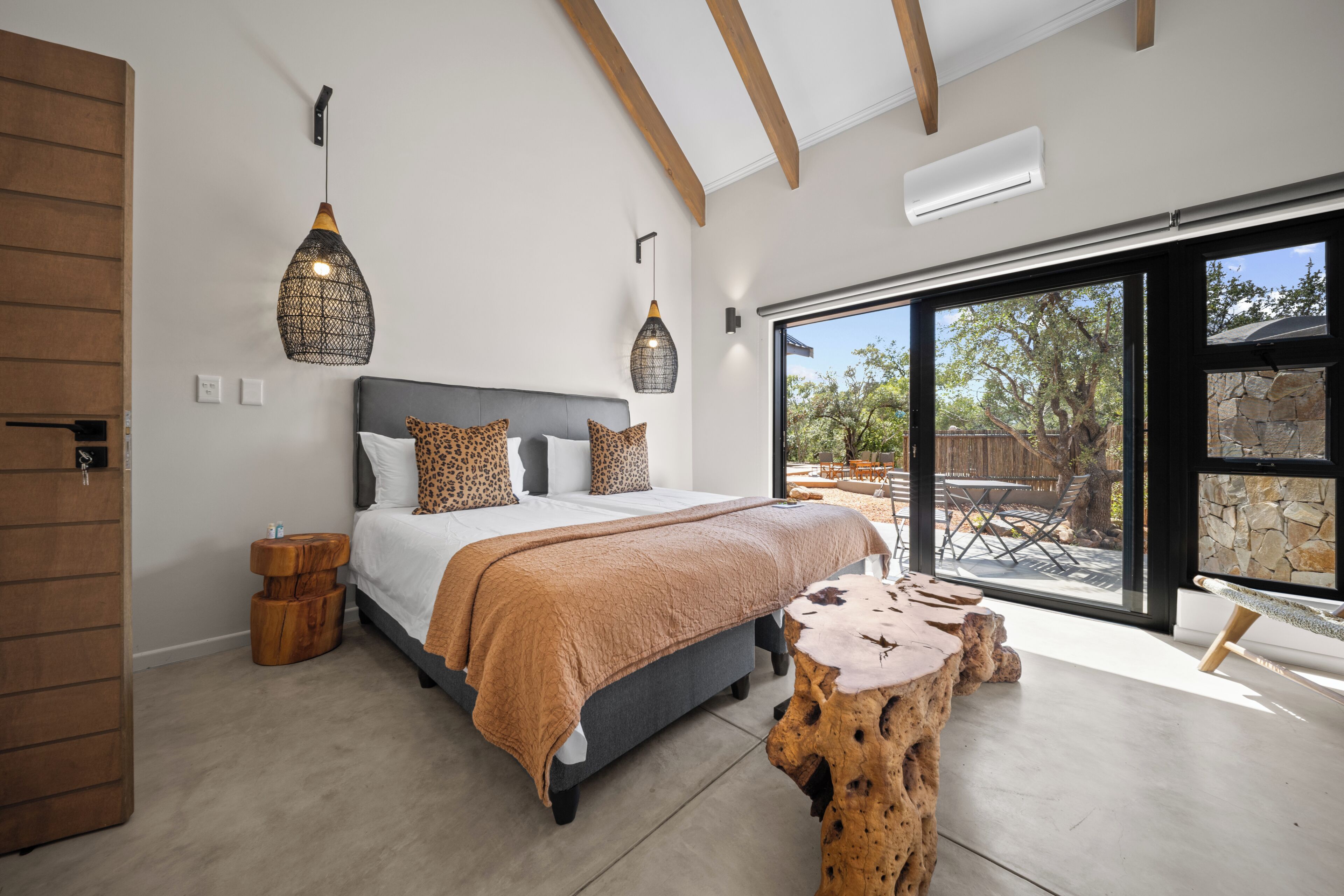 A bedroom with a large bed, a wooden headboard, a hanging light fixture, a bench, and a view of the outdoors.