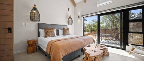 A bedroom with a large bed, a wooden headboard, a hanging light fixture, a bench, and a view of the outdoors.