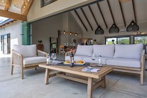En rummelig udendørs siddeplads med et træbord, to sofaer og et spisebord dækket med tallerkener, glas og en flaske.
