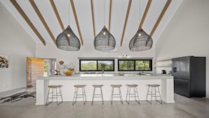 A modern kitchen with a central island, bar stools, pendant lighting, and a black refrigerator. - Kimbilia Luxury Villa  (Hoedspruit)