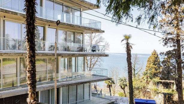 En moderne bygning med flere etager, store glasvinduer, en balkon og udsigt til havet.