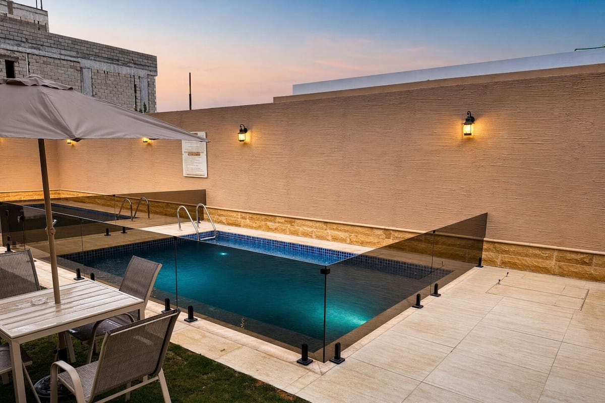 An outdoor swimming pool with a glass barrier, a table with chairs, and a grassy area.