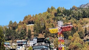 Cielo azul despejado con un parapente a lo lejos, una torre roja y amarilla, y una ladera con edificios.