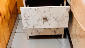 Una mesita de noche pequeña y blanca con cajones, un enchufe y una cama con cabecero blanco.