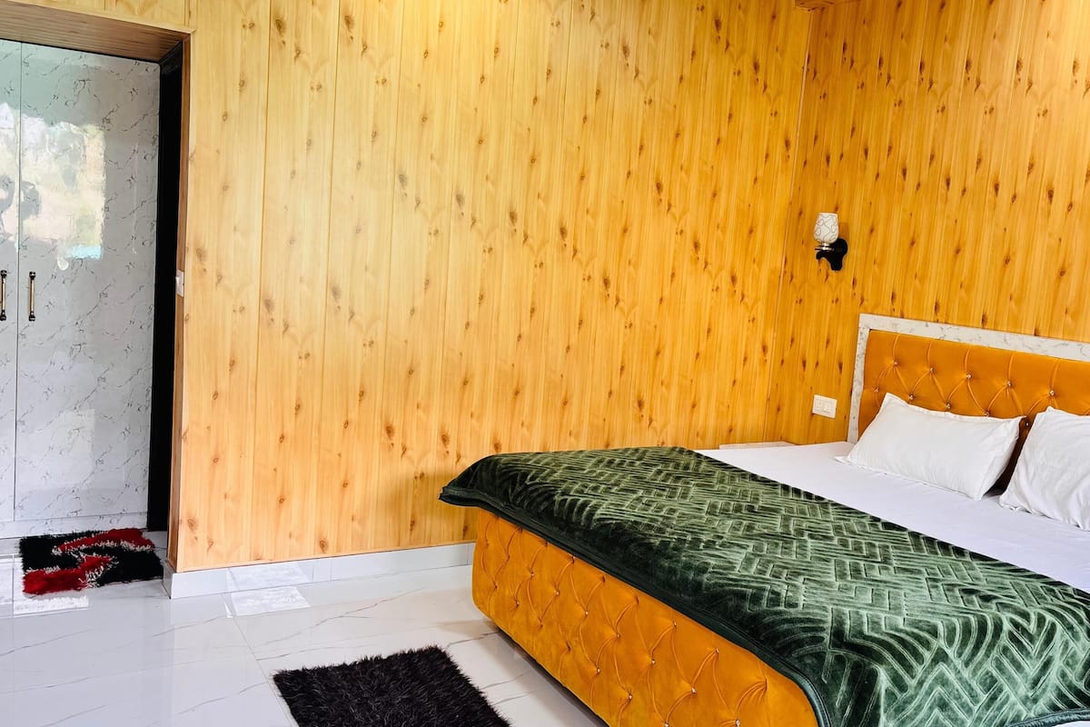 A bedroom with a wooden wall, a bed with a green patterned bedspread, white pillows, and a black rug.