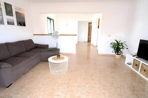 A living room with a brown sofa, a round coffee table with a plant, a white fan, and a TV on a white cabinet.