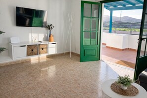 A room with a TV mounted on the wall, a white console table with storage, a green door leading outside, and a view of a mountainous landscape through the door.